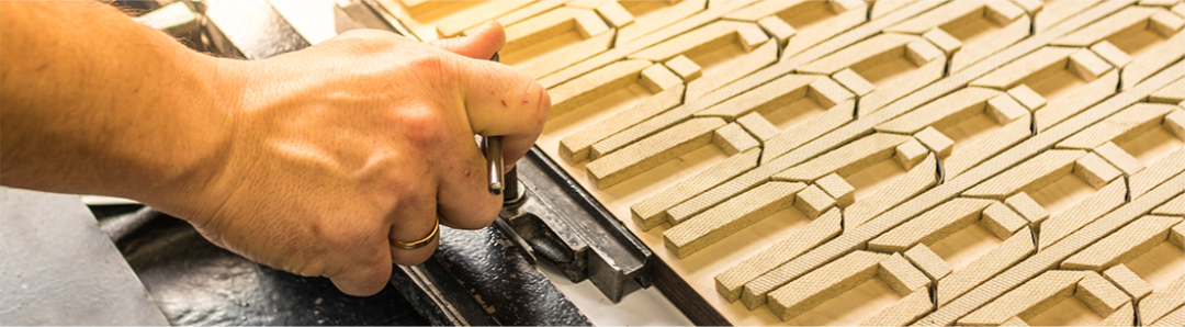 A close-up of a persons hand adjusting or assembling a patterned, interlocking tile or panel, possibly during a manufacturing or crafting process. The pattern features repeating geometric shapes.