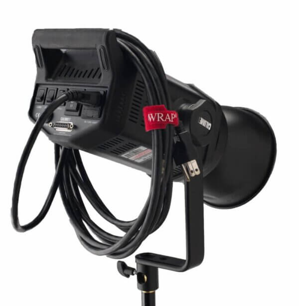 A professional studio light with controls and ports visible on the back, a coiled power cable secured with a red strap, and mounted on a black stand against a white background.