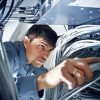 A man in a server room carefully inspects and adjusts network cables connected to equipment, surrounded by numerous wires and electronic devices.