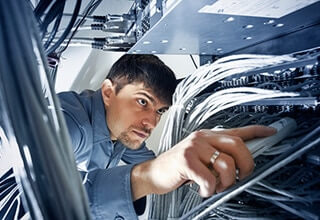 A man in a server room carefully inspects and adjusts network cables connected to equipment, surrounded by numerous wires and electronic devices.