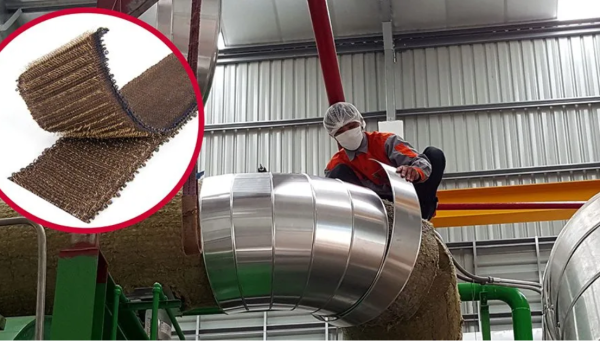 A worker in safety gear wraps VELCRO® Brand HI-GARDE® Fasteners around a large industrial pipe; an inset shows a close-up of the brown, fibrous fastener. The factory setting features metal pipes and a metal roof.