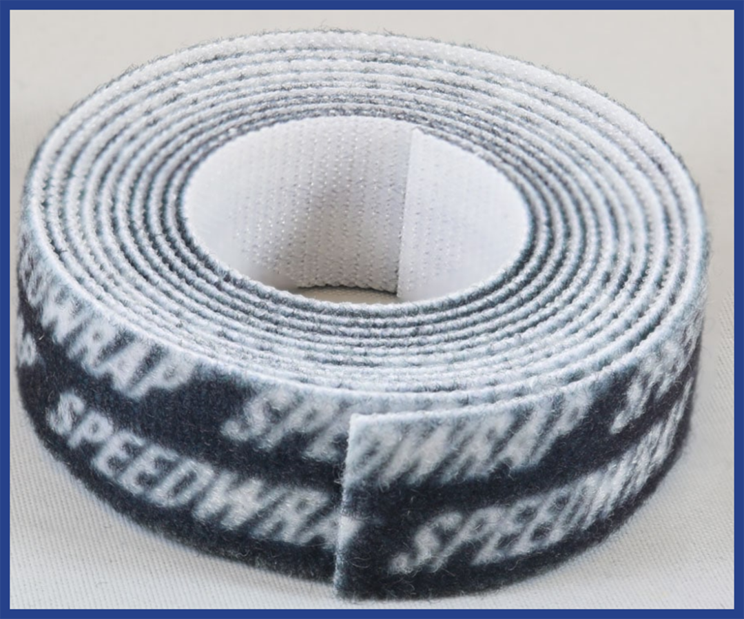 A close-up of a coiled black and white Velcro cable tie with the word SPEEDWRAP repeatedly printed in white on the black side. The tie is placed on a light surface with a blue border around the image.